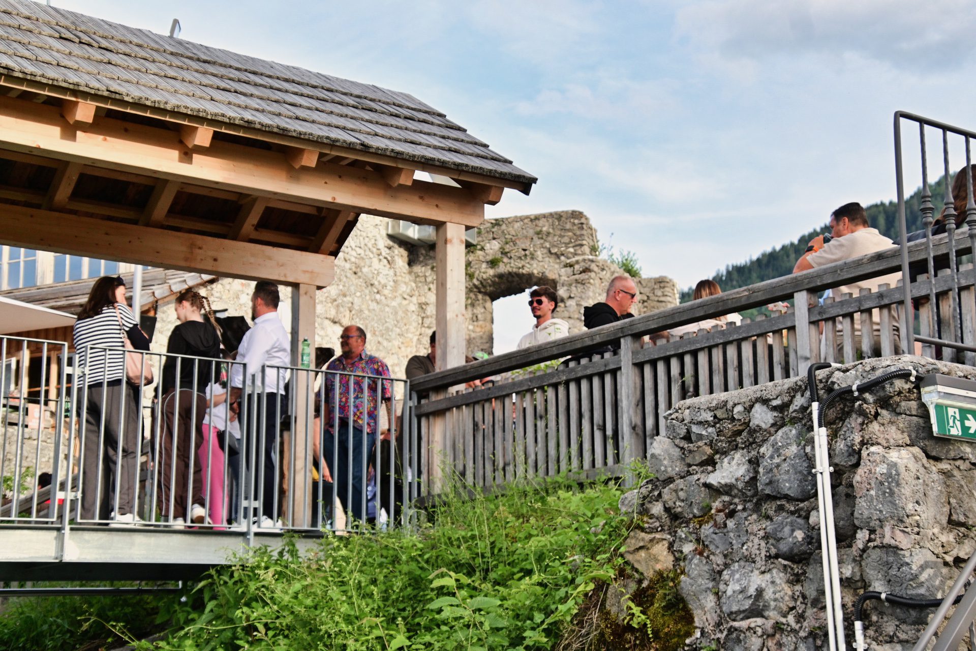 Menschen stehen auf einer Aussichtsplattform vor alten Steinruinen und einem Holzgebäude