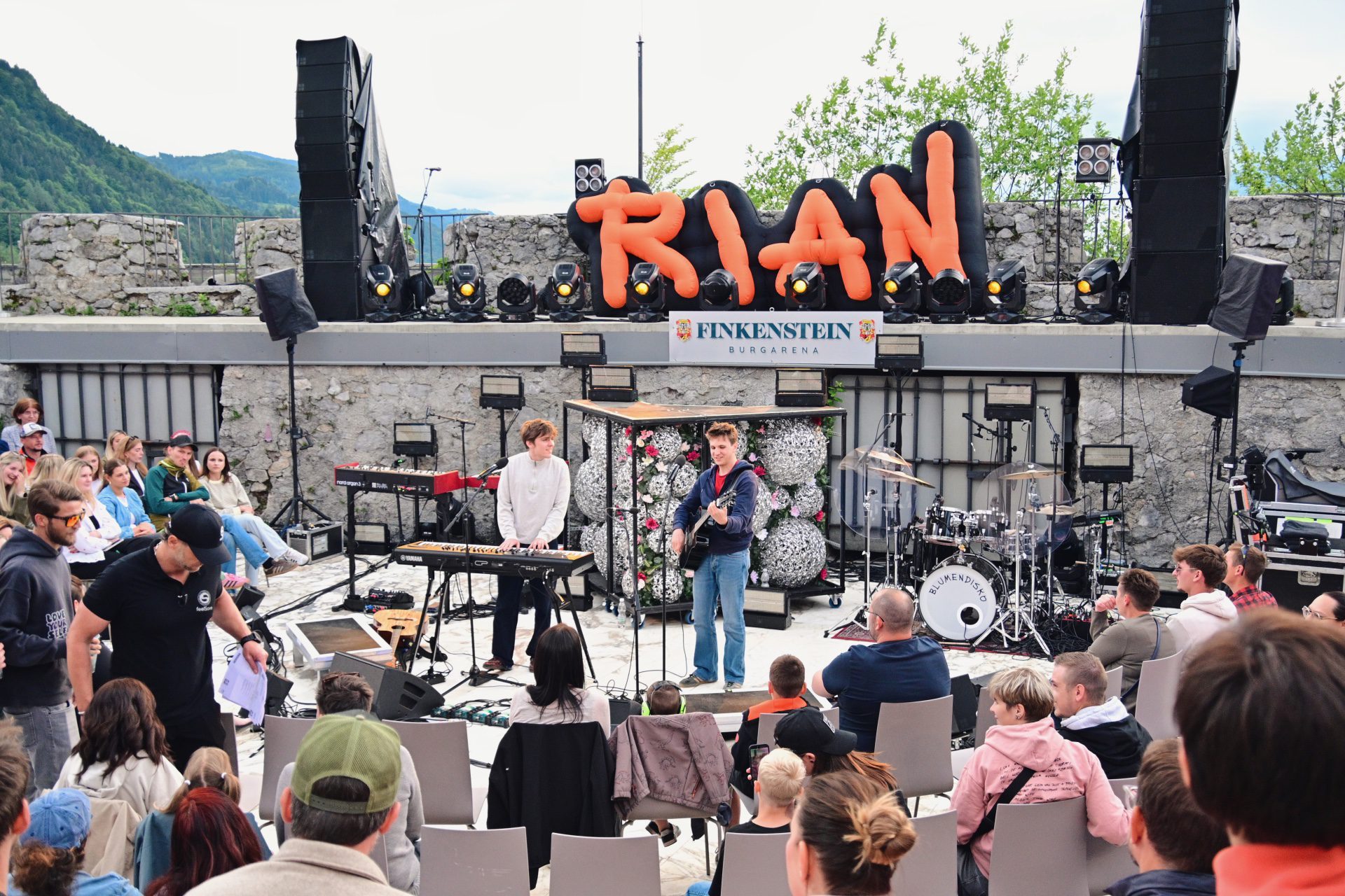 Zwei Musiker auf Bühne mit großem orangem RIAN-Schriftzug und sitzendem Publikum im Freien