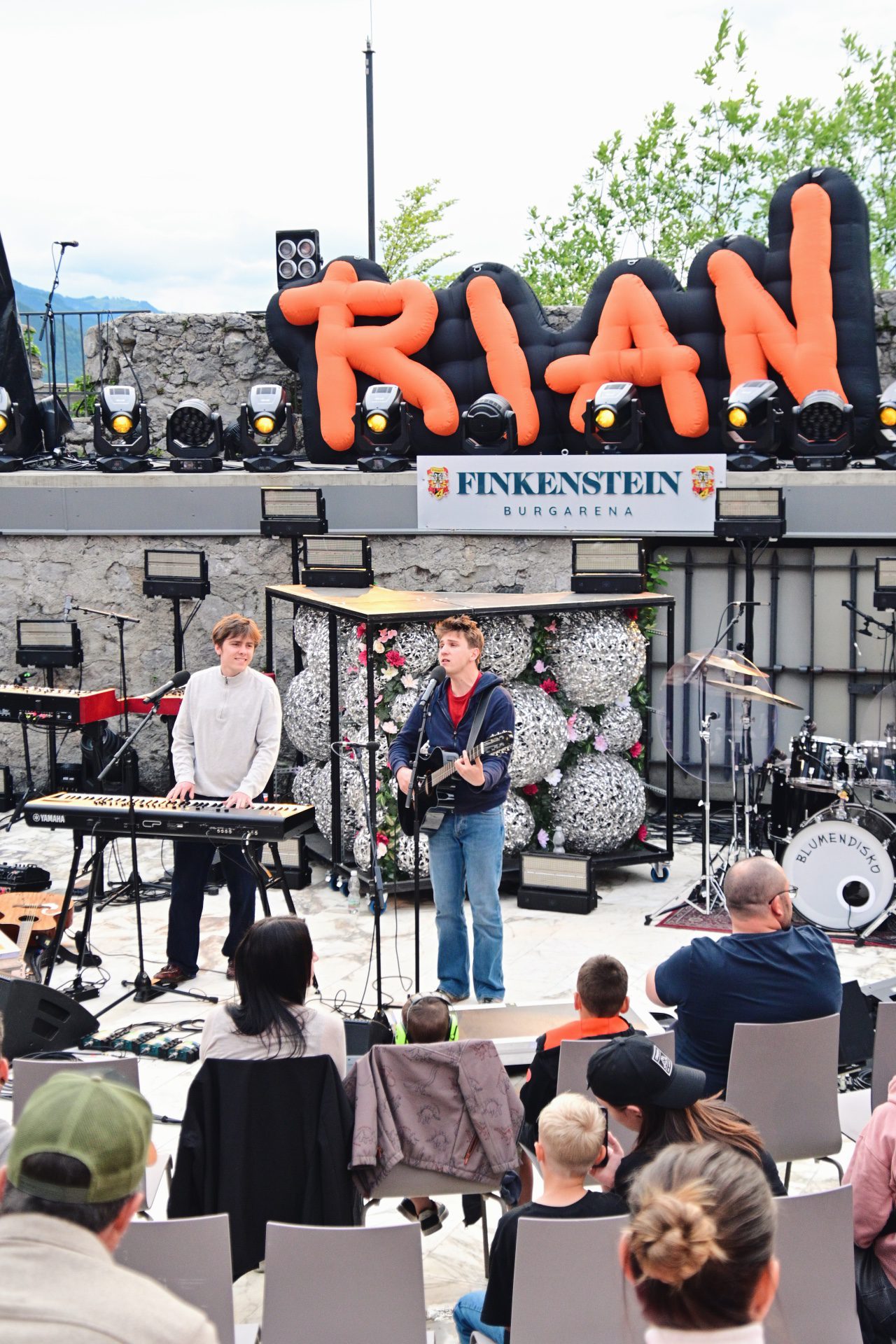 Zwei Musiker mit Keyboard und Gitarre auf Bühne vor orangen RIAN-Buchstaben und Publikum.