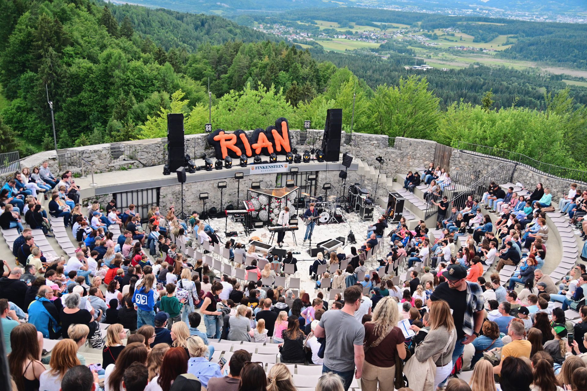 Zuschauer in einem Amphitheater bei einem Konzert mit Bühne und Band vor grüner Landschaft