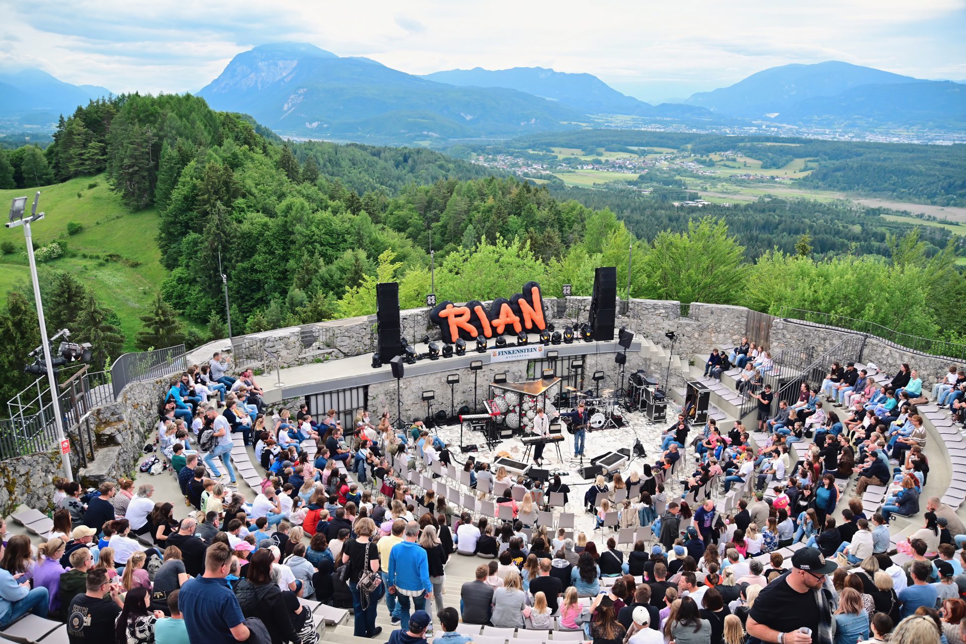 Großes Publikum auf Steinstufen vor Bühne mit orangem RIAN-Schriftzug und Bergen im Hintergrund
