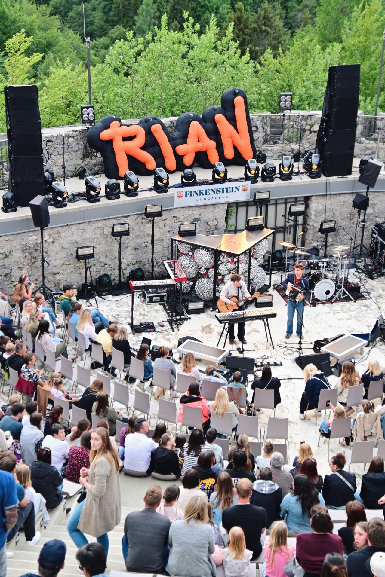 Zwei Musiker spielen Gitarre und Akkordeon auf einer Bühne vor sitzendem Publikum mit RIAN-Schriftzug im Hintergrund