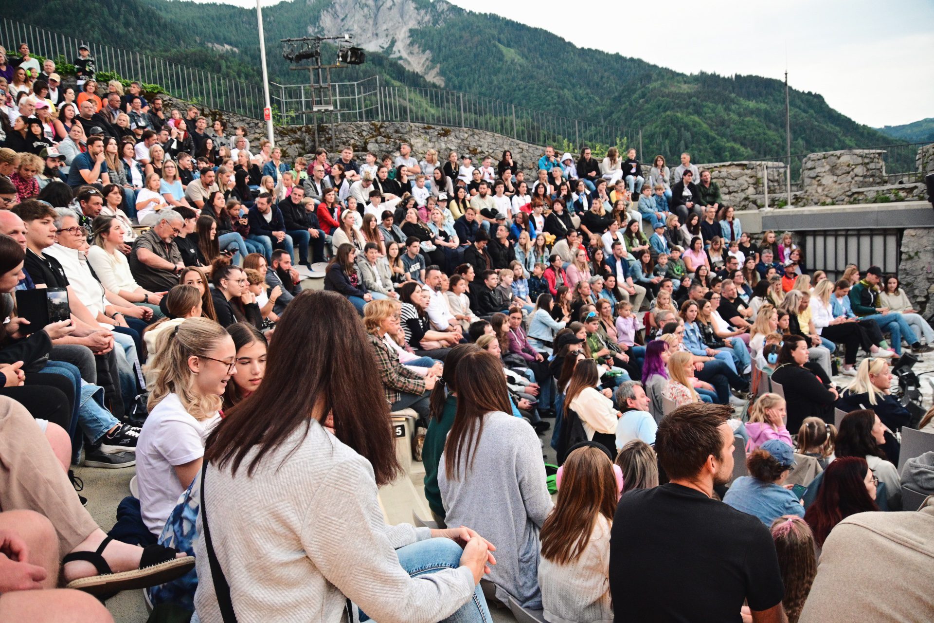 Zahlreiche Zuschauer sitzen auf Stufen eines Freilufttheaters vor bewaldeten Bergen