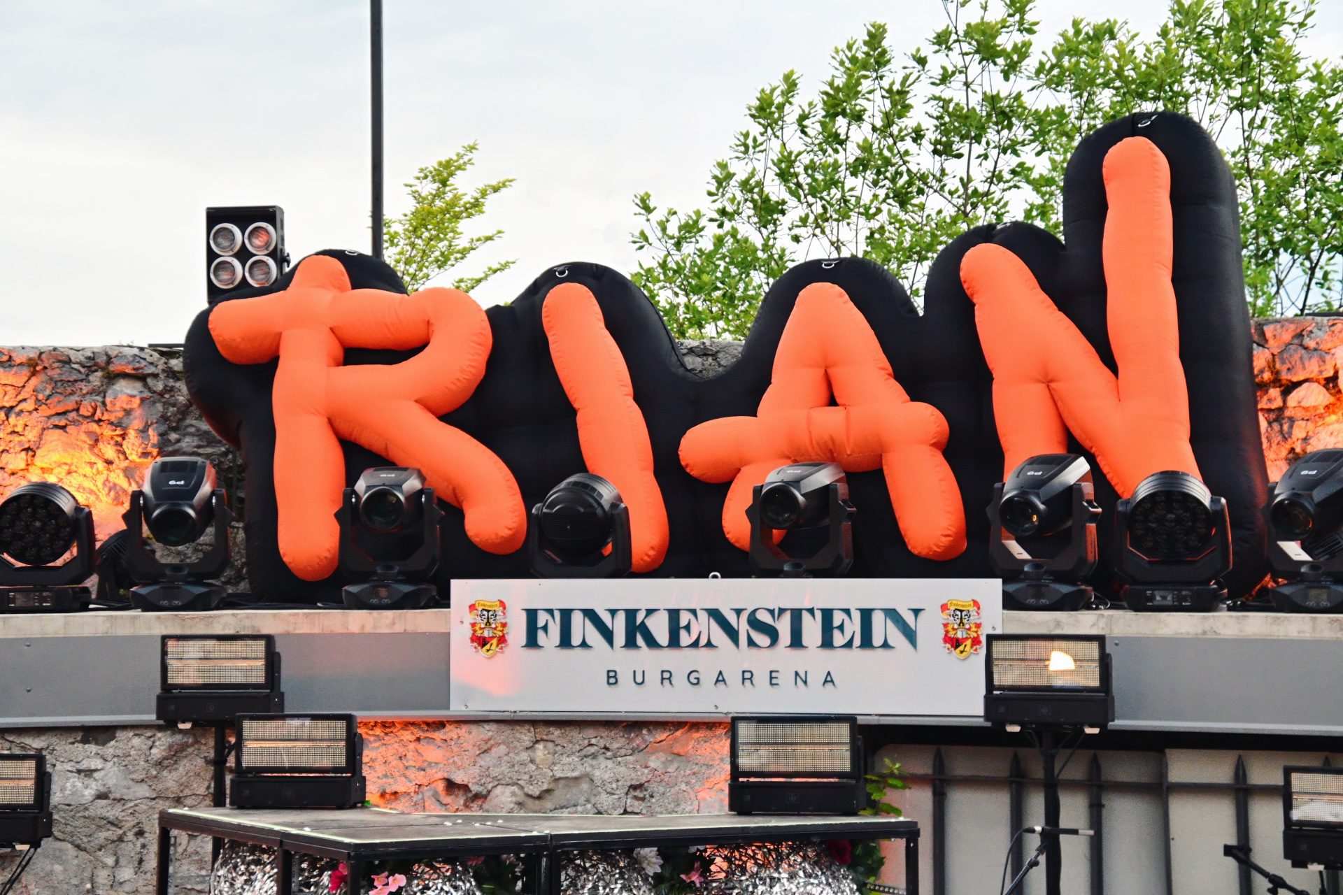 Aufblasbare orangene Buchstaben formen das Wort RIAN vor einer Steinwand mit Bühnenbeleuchtung