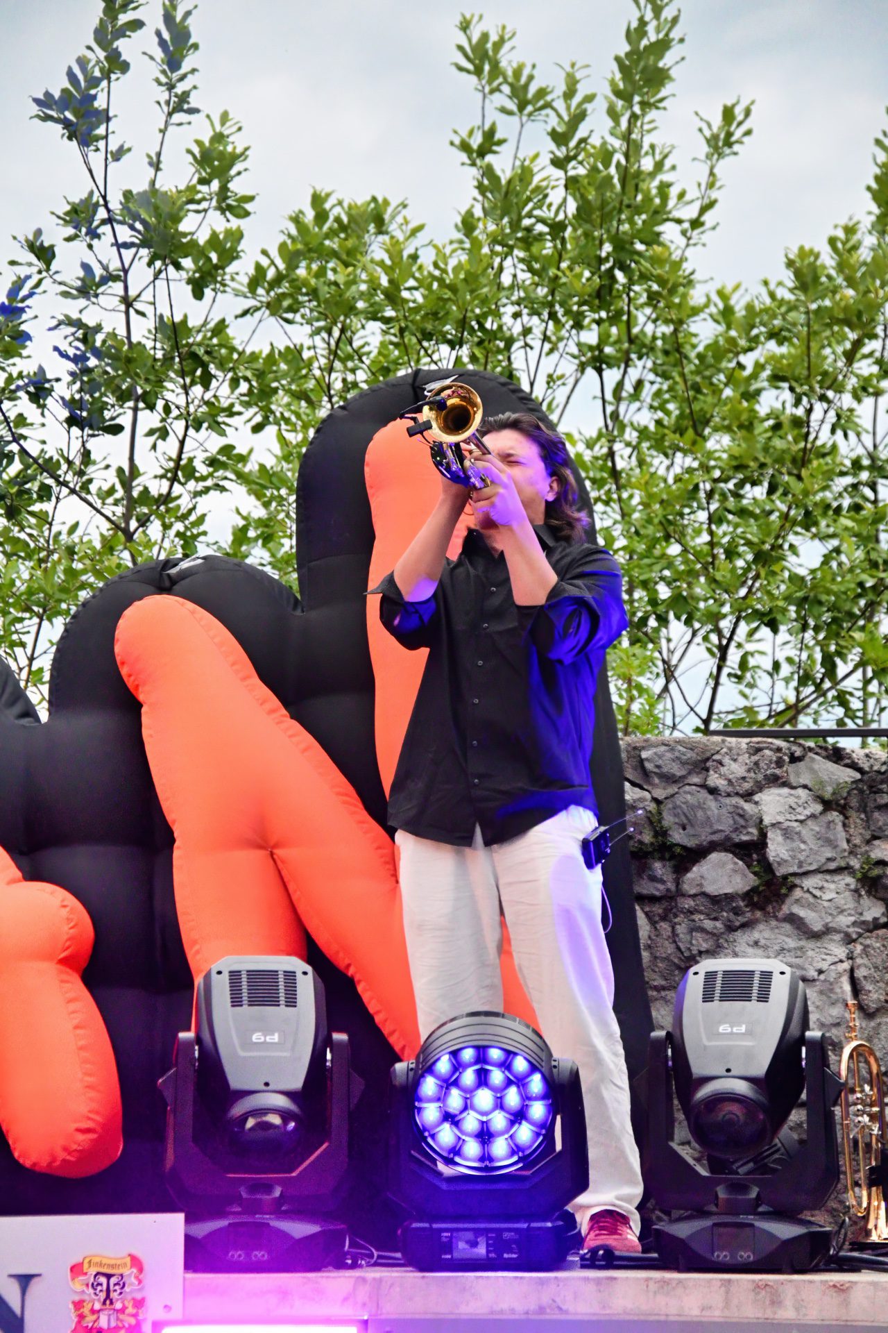 Ein Musiker mit langen Haaren spielt Trompete vor einer großen aufblasbaren Skulptur auf der Bühne.