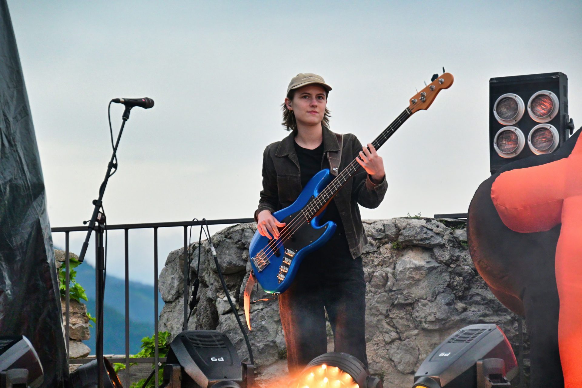 Musikerin spielt blaue E-Bassgitarre auf einer Freilichtbuehne mit Steinmauer und Scheinwerfern