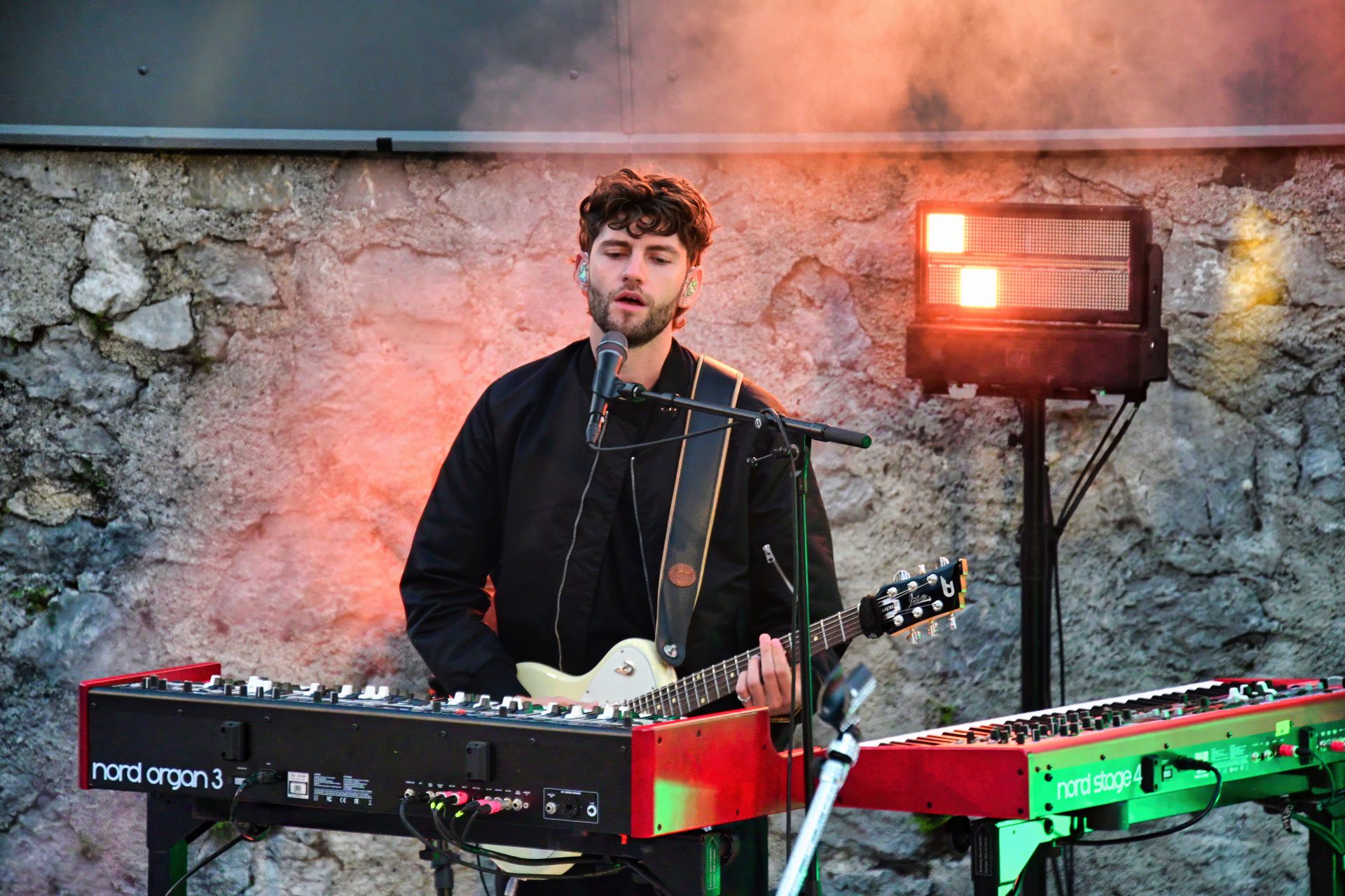 Ein bärtiger Musiker mit lockigem Haar spielt E-Gitarre hinter Keyboards vor einer Steinwand.