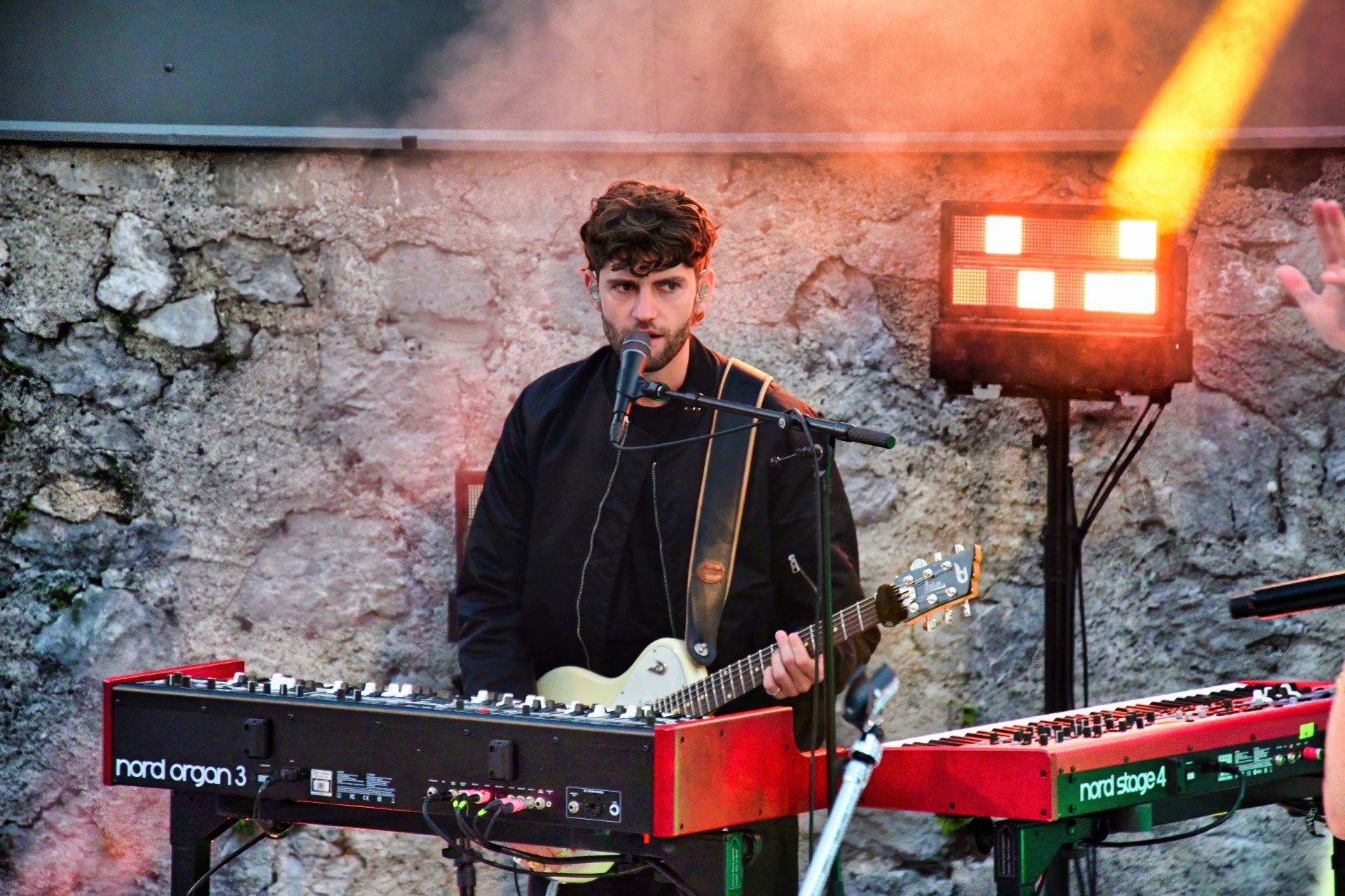 Bärtiger Musiker mit dunklen Locken spielt E-Gitarre auf Bühne vor Steinwand mit Keyboards und Scheinwerfern
