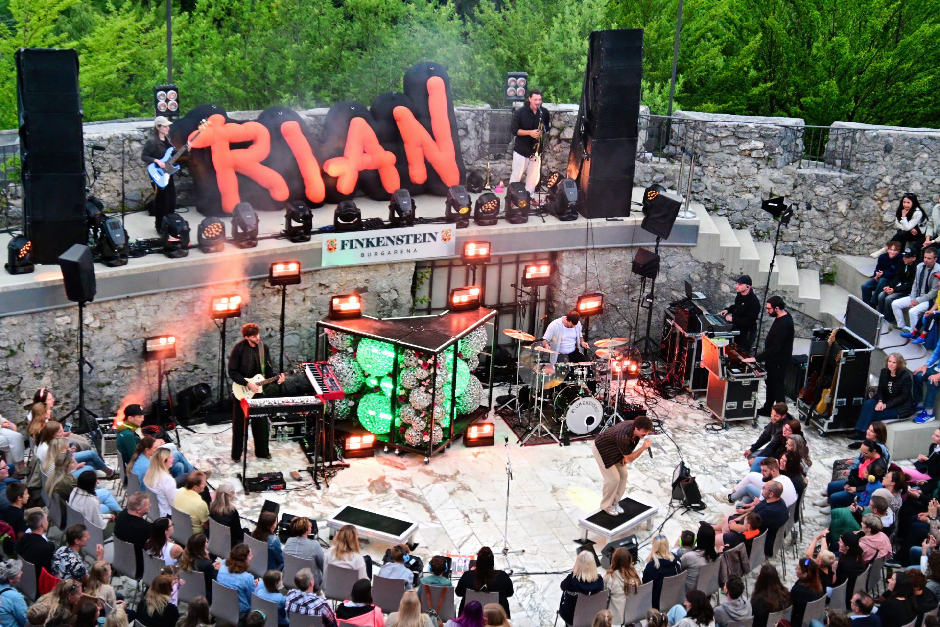 Bandmitglieder spielen auf einer beleuchteten Bühne mit RIAN-Schriftzug vor Publikum in Finkenstein