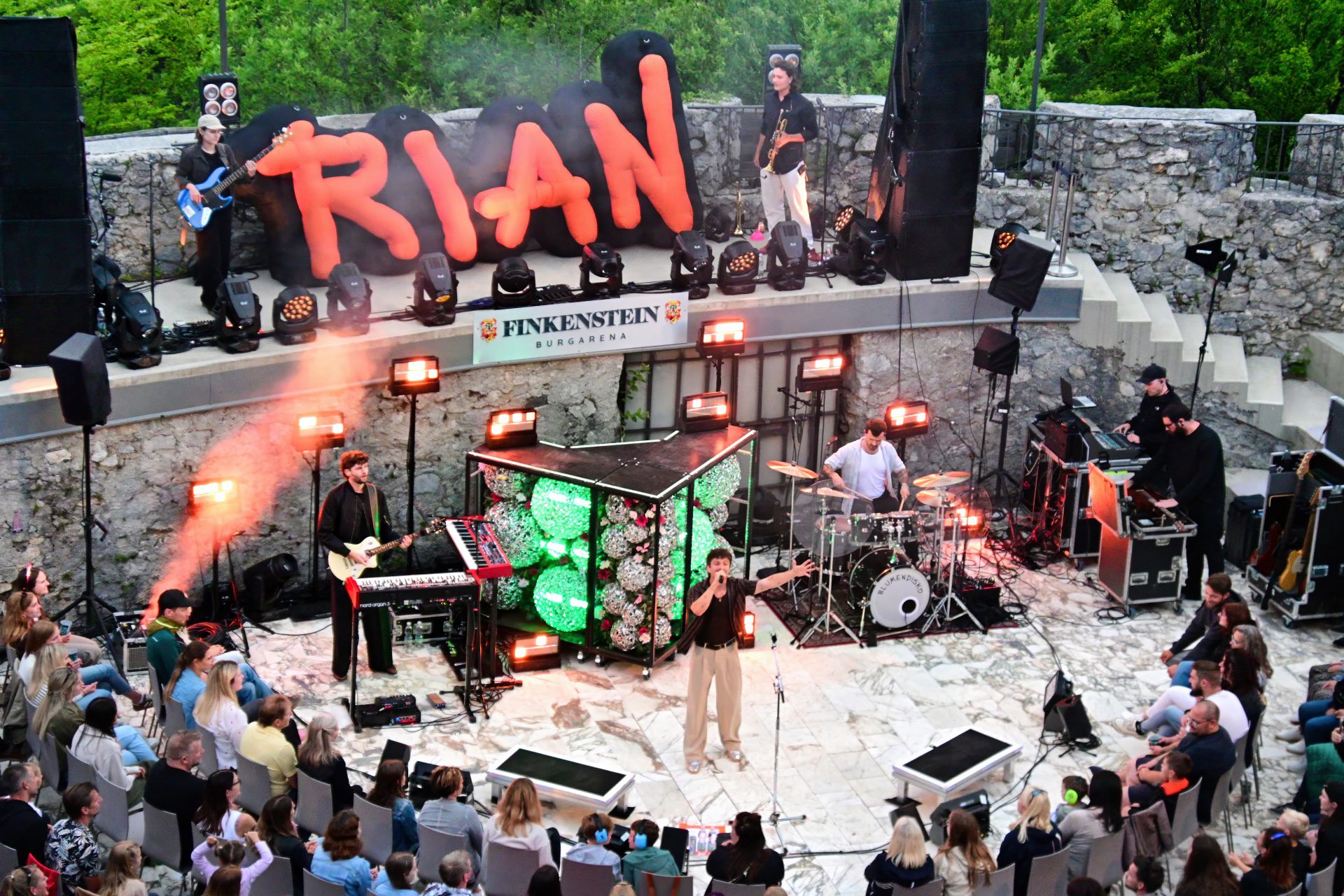 Sänger mit ausgestreckten Armen auf der Bühne vor sitzendem Publikum bei einem Konzert in der Burgarena Finkenstein
