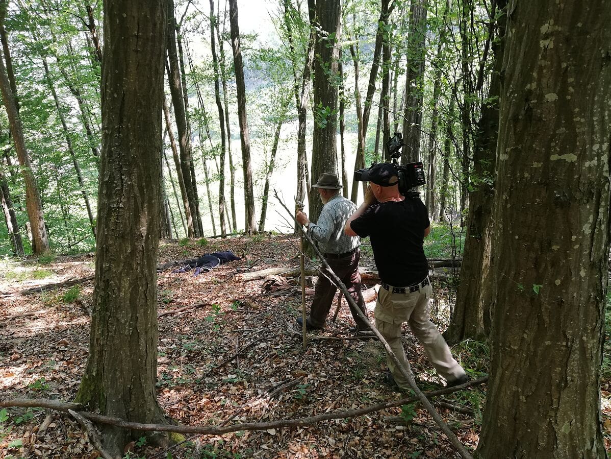Kameramann filmt einen Mann mit Hut im Wald neben einer abgedeckten Leiche