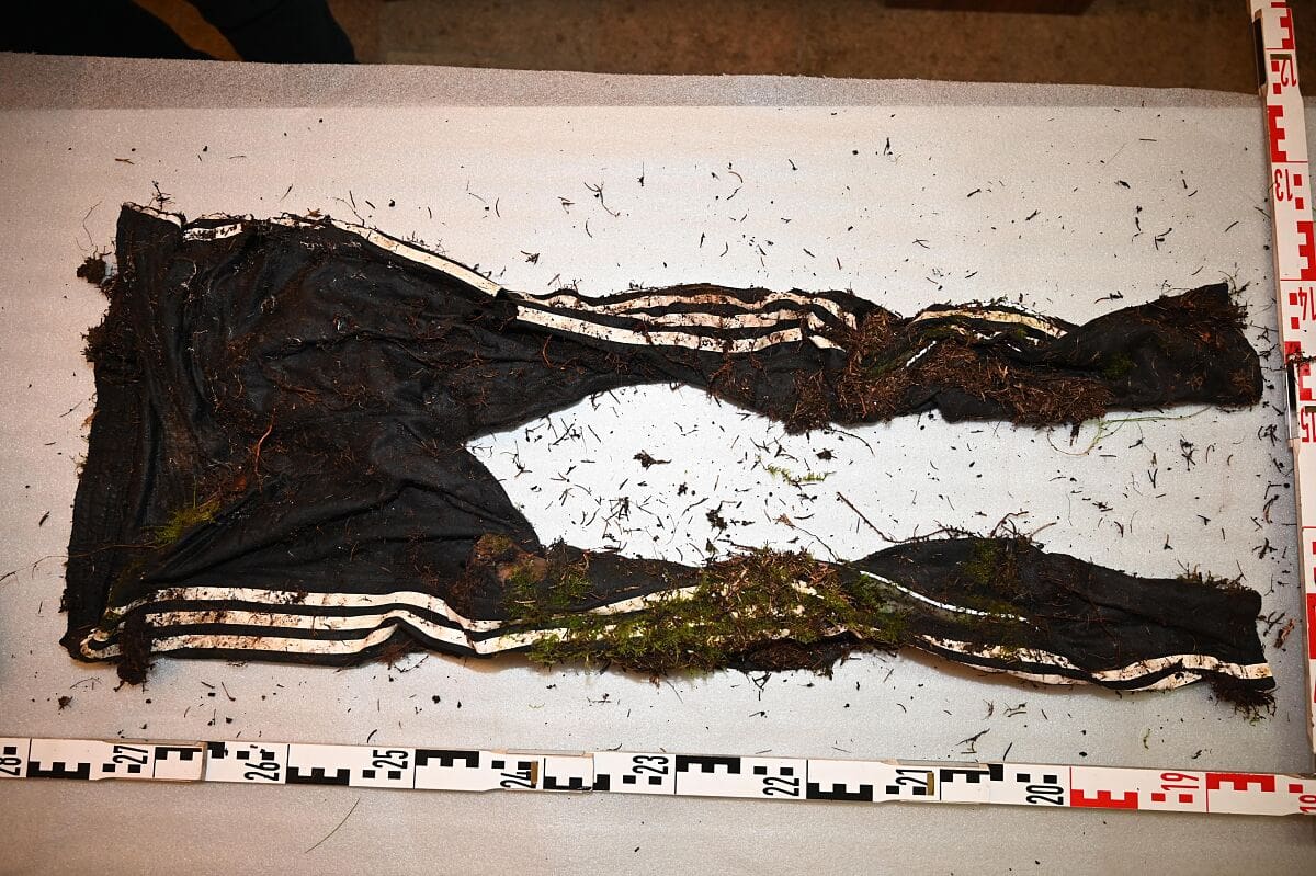 Schwarze lange Hose mit weißen Streifen, bedeckt mit Erde, Wurzeln und Moos auf weißer Unterlage mit Maßband