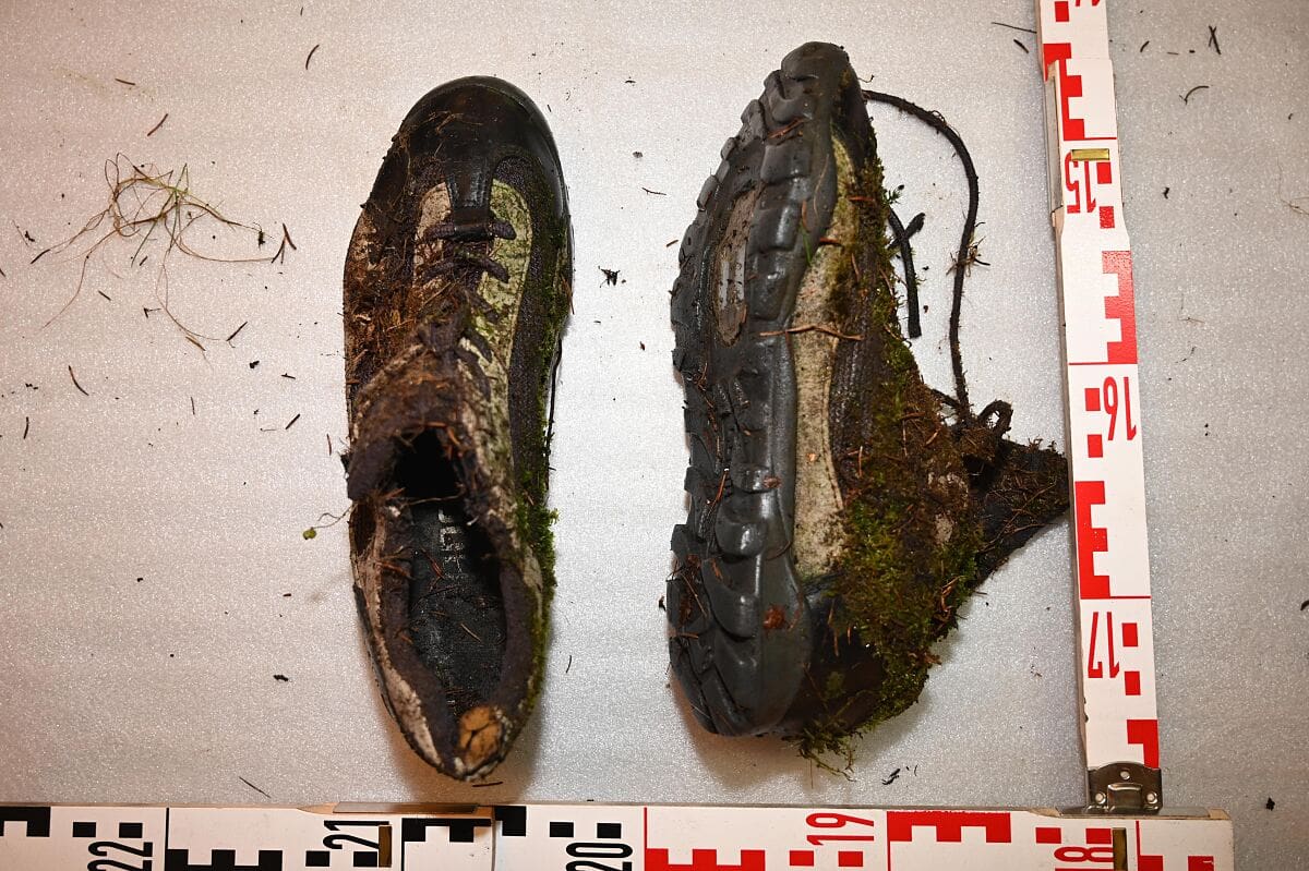 Zwei stark verschmutzte und moosbewachsene Wanderschuhe neben Messskalen auf weißer Unterlage.