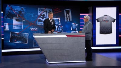 Zwei Moderatoren stehen in einem Fernsehstudio vor einer Ermittlungstafel mit Fahrrad und Beweisfotos