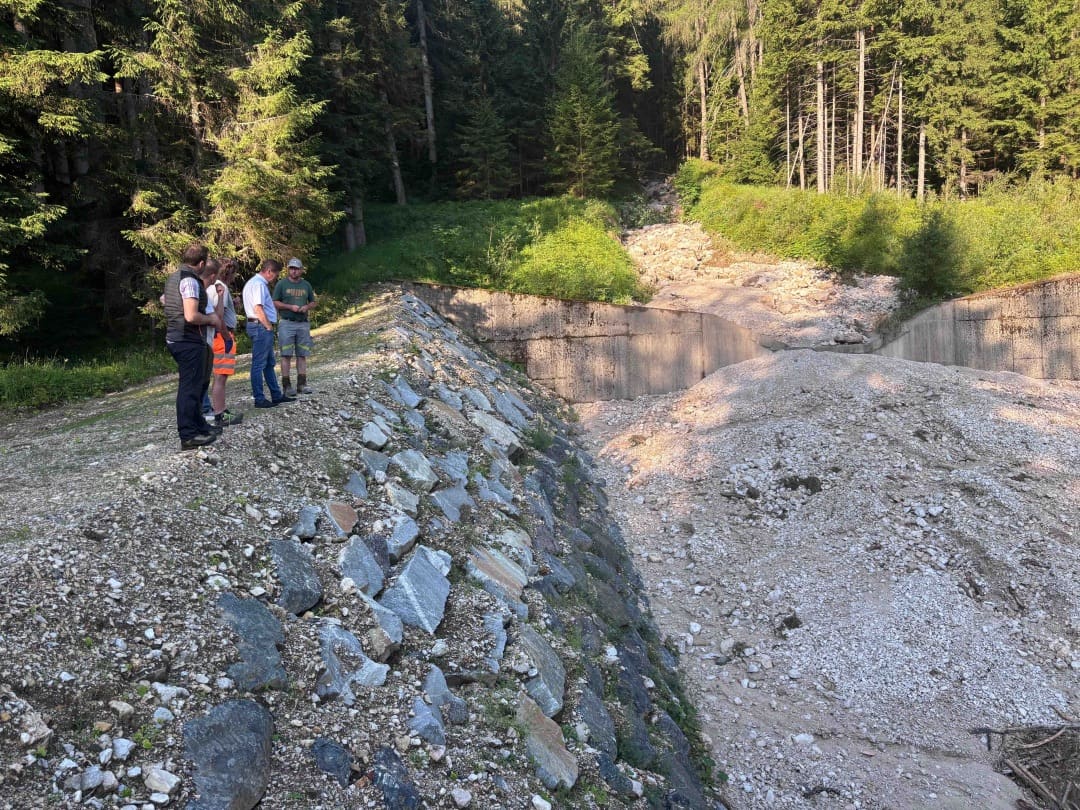 Gruppe von Personen steht neben einem Betonrückhaltebecken in einem schlammbedeckten Bachbett