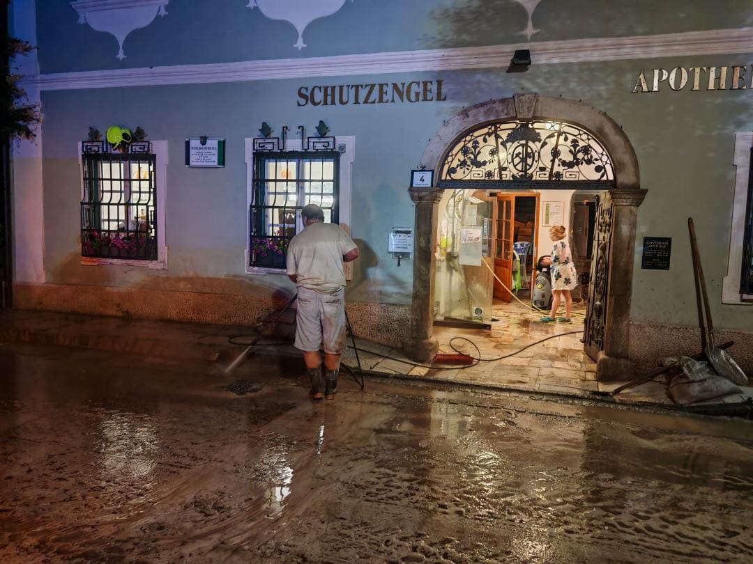 Ein Mann reinigt mit einem Hochdruckreiniger eine schlammbedeckte Straße vor einer Apotheke bei Nacht