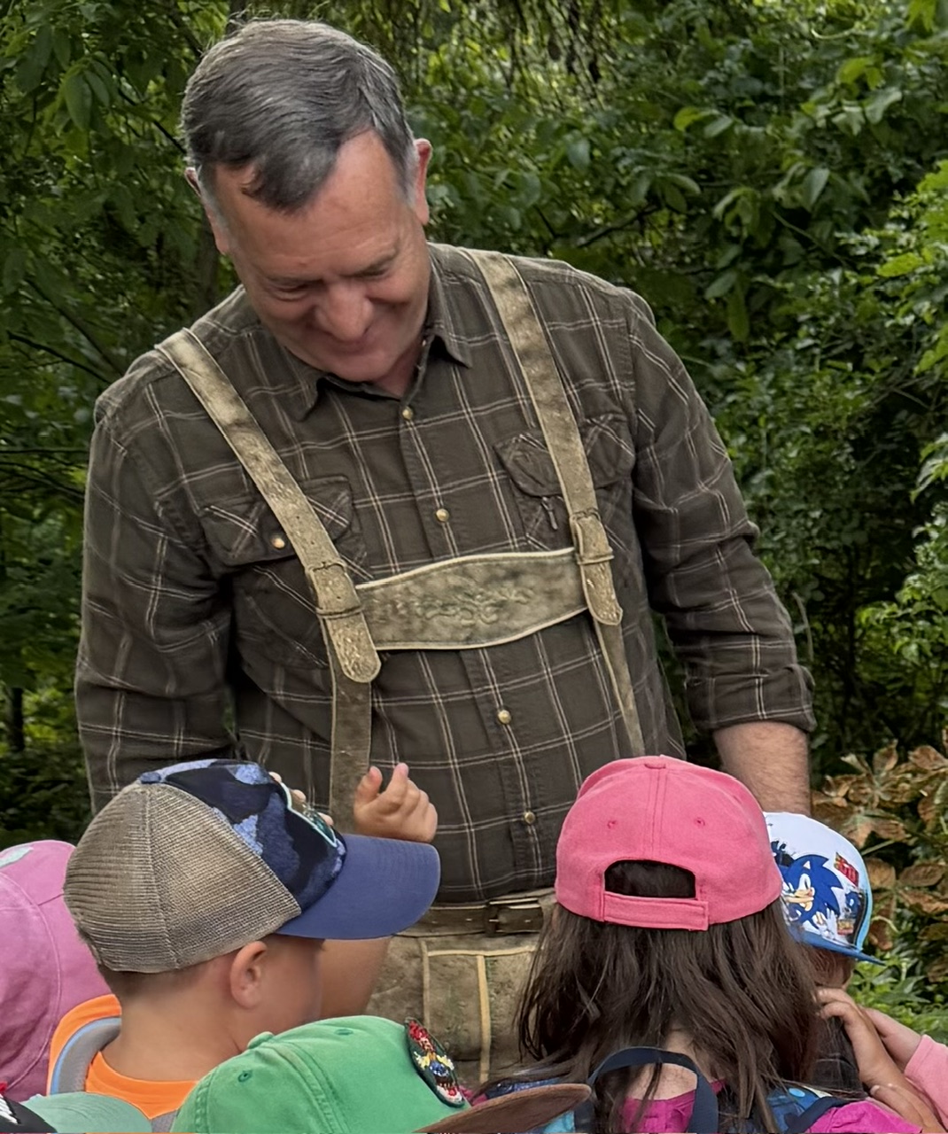 Ein Mann in kariertem Hemd und Tracht spricht mit Kindergartenkindern im Wald