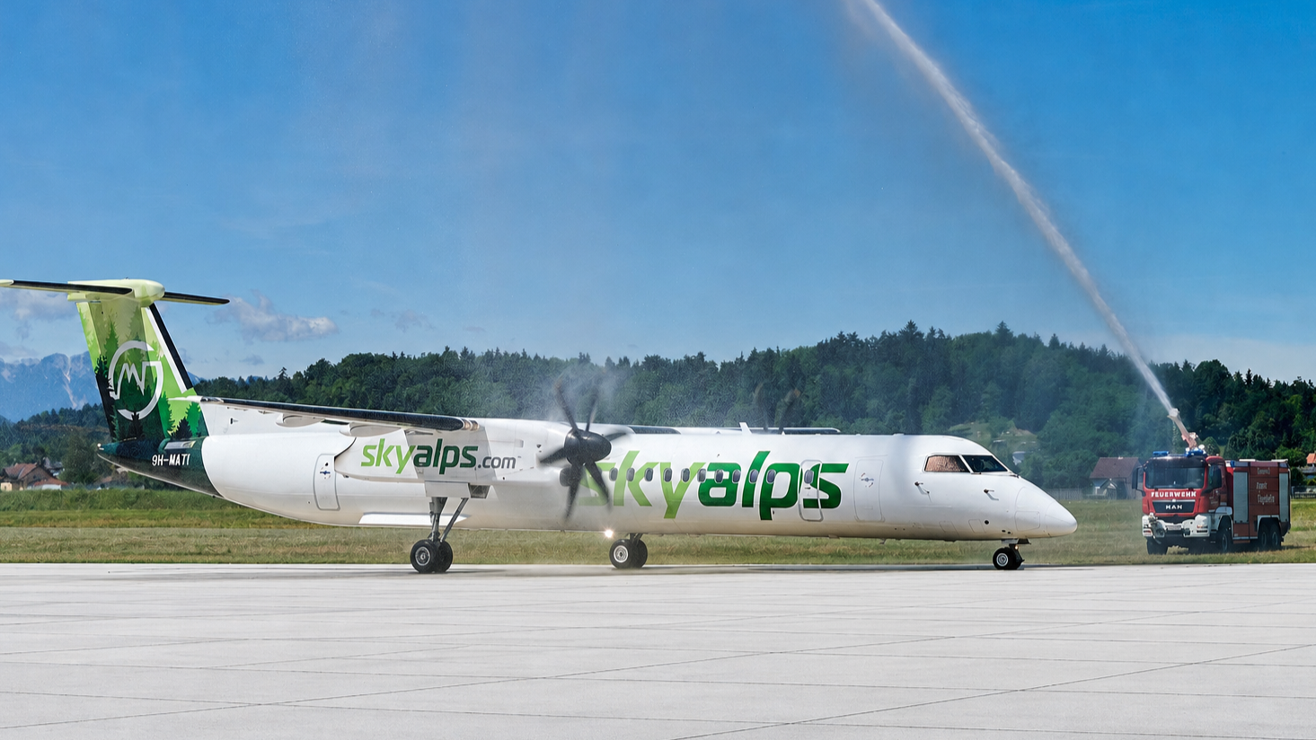 Weißes SkyAlps-Propellerflugzeug auf dem Vorfeld während Wassersalut durch Feuerwehrfahrzeug