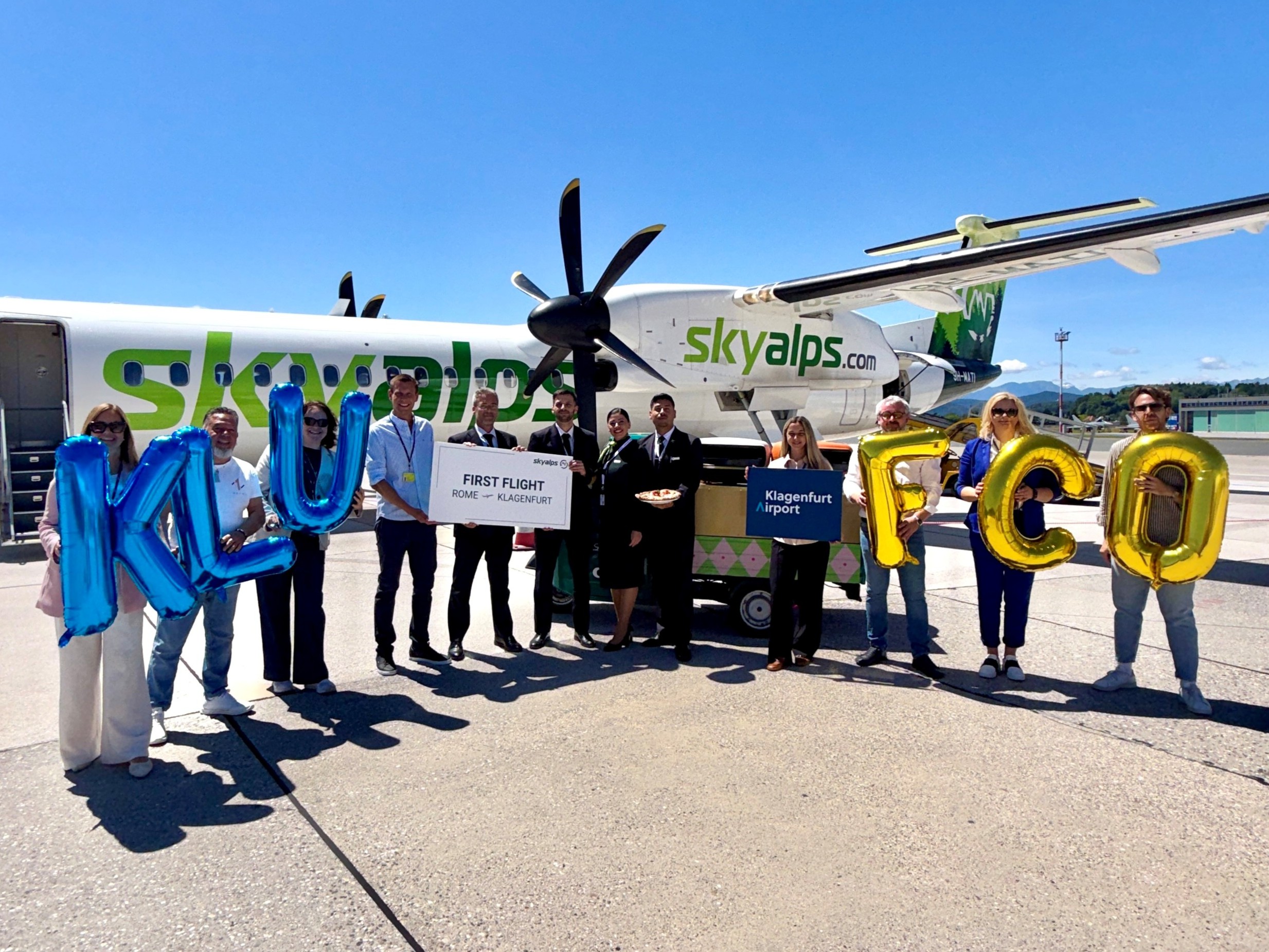 Gruppe von Menschen mit Luftballons KLU und FCO vor einem SkyAlps Flugzeug am Flughafen