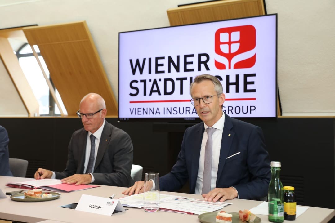 Zwei Männer in Anzügen sitzen an einem Konferenztisch vor einem Bildschirm mit dem Logo der Wiener Städtischen.