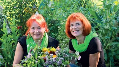 Zwei Frauen mit roten Haaren sitzen in einem Garten und halten einen Korb mit Blumen.