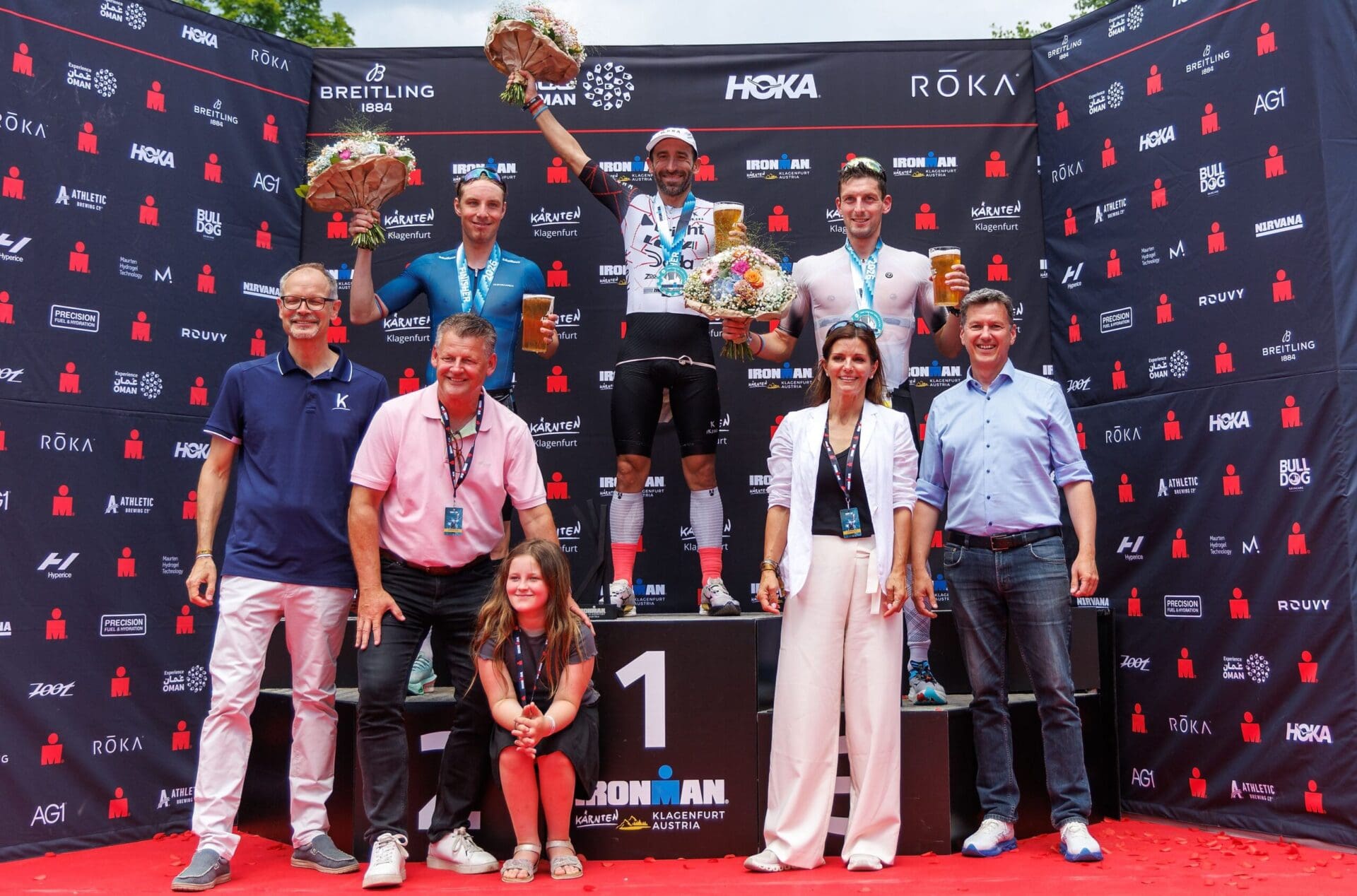 Mattia Ceccarelli steht als Sieger auf dem Podium des Ironman Austria in Klagenfurt und hält einen Blumenstrauß hoch.