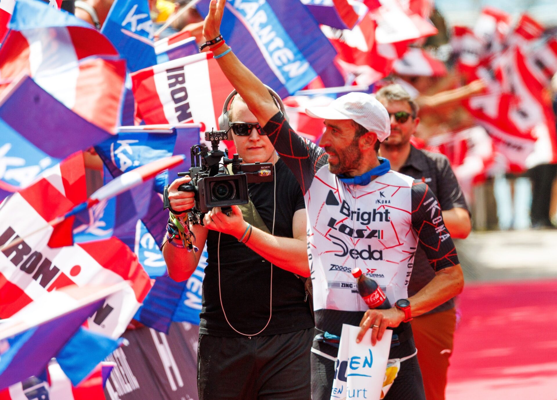 Triathlet Mattia Ceccarelli neben einem Kameramann mit Headset vor bunten Ironman Flaggen am Ziel