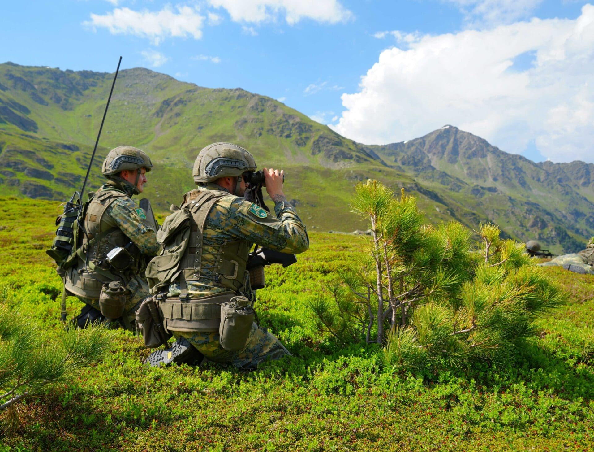 Zwei Soldaten in Tarnuniform knien in bergiger Landschaft, einer blickt durch Fernglas