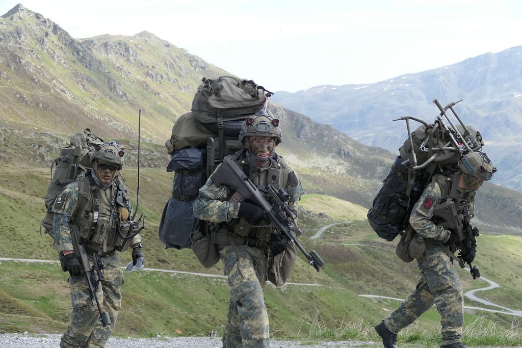 Drei Soldaten in Tarnuniform mit schweren Rucksäcken und Waffen marschieren durch eine Gebirgslandschaft.