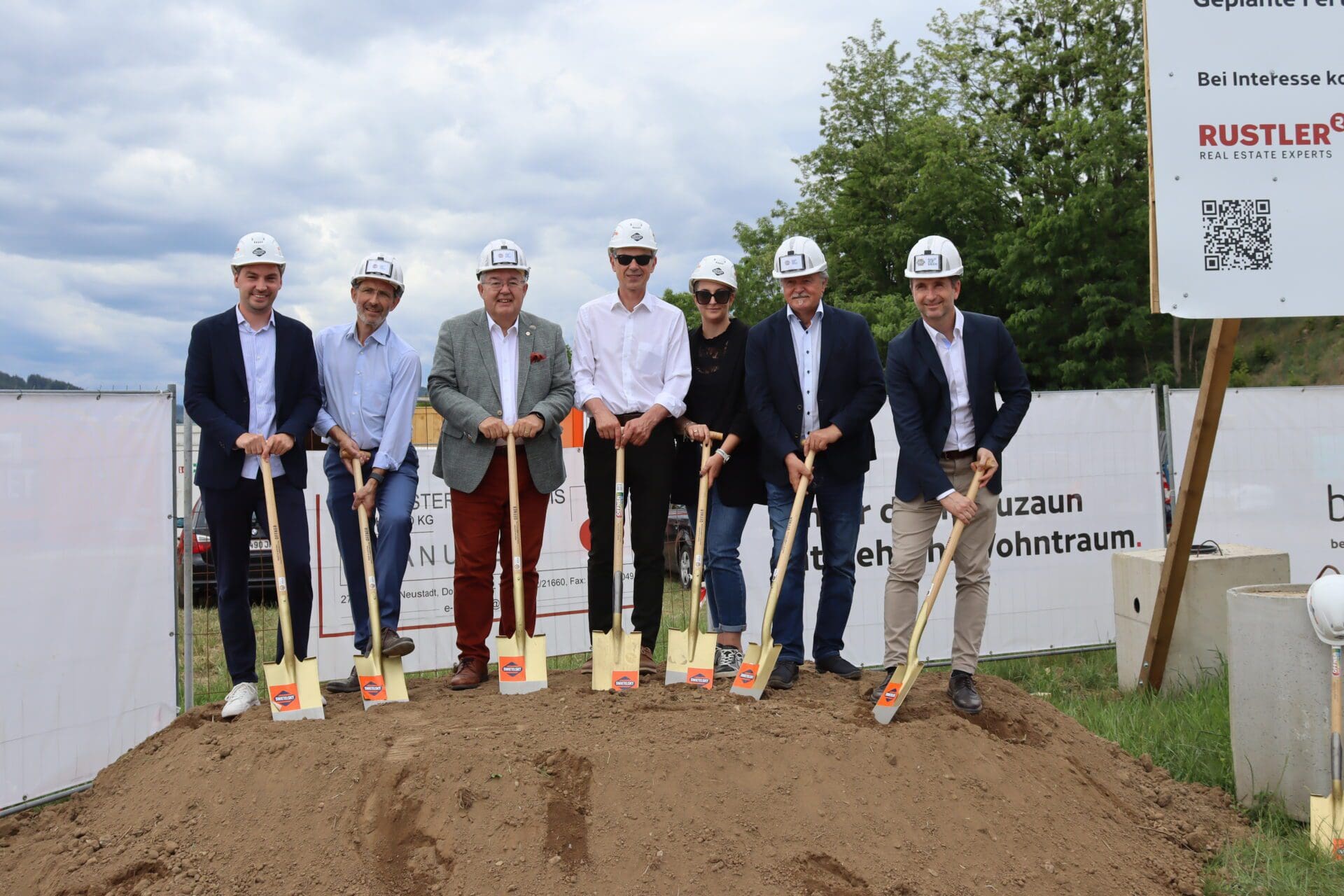 Gruppe von Personen in Anzügen und Schutzhelmen mit goldenen Spaten auf einem Erdhügel bei einer Baustelle