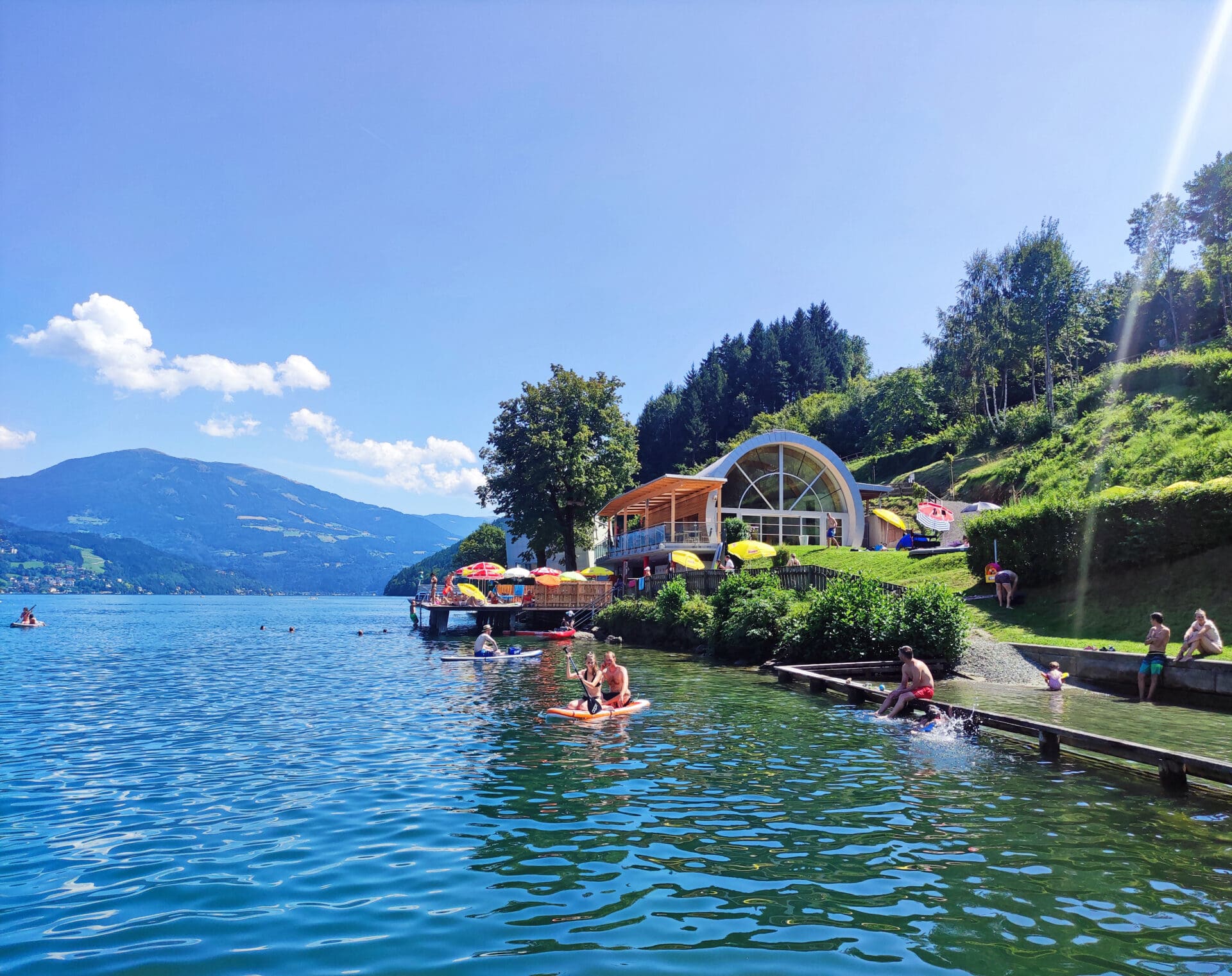 Menschen auf einem orangefarbenen Paddleboard vor einem modernen Gebäude am Seeufer