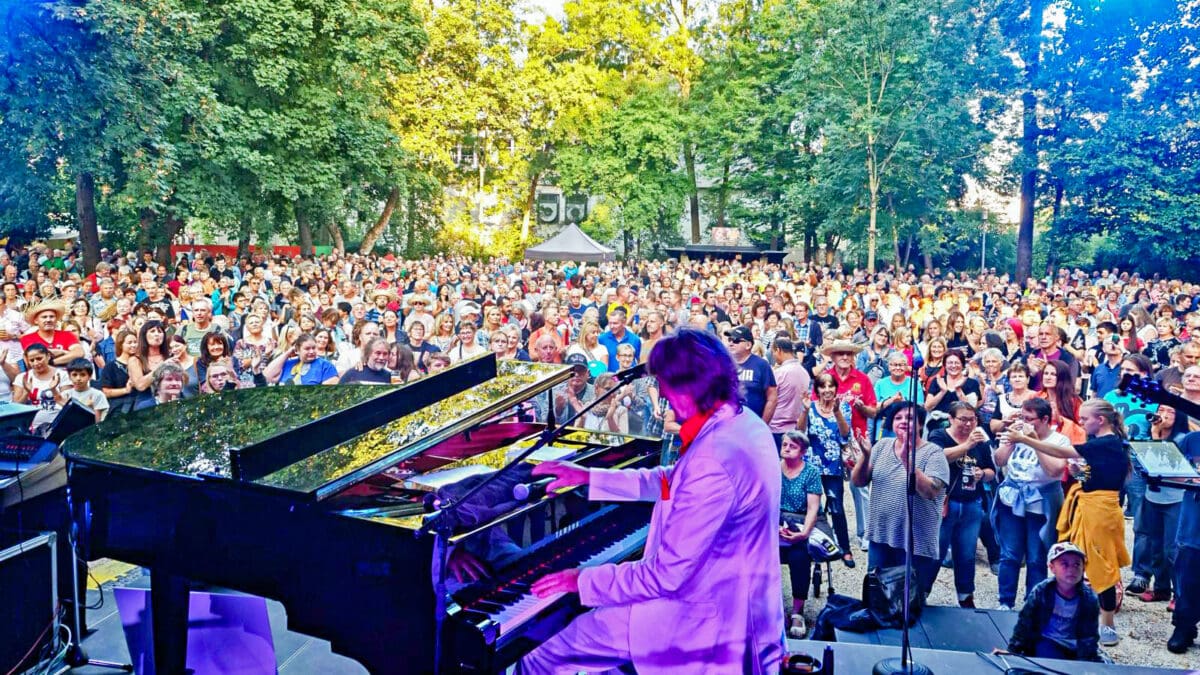 Ein Pianist in einem hellen Anzug spielt auf einem Flügel vor einer großen Menschenmenge im Park.