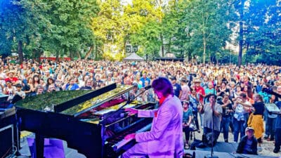 Ein Pianist in einem hellen Anzug spielt auf einem Flügel vor einer großen Menschenmenge im Park.