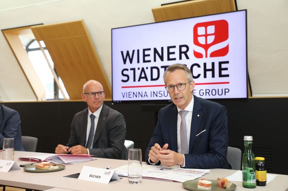 Zwei Männer in Anzügen sitzen an einem Tisch vor einem Bildschirm mit dem Logo der Wiener Städtischen