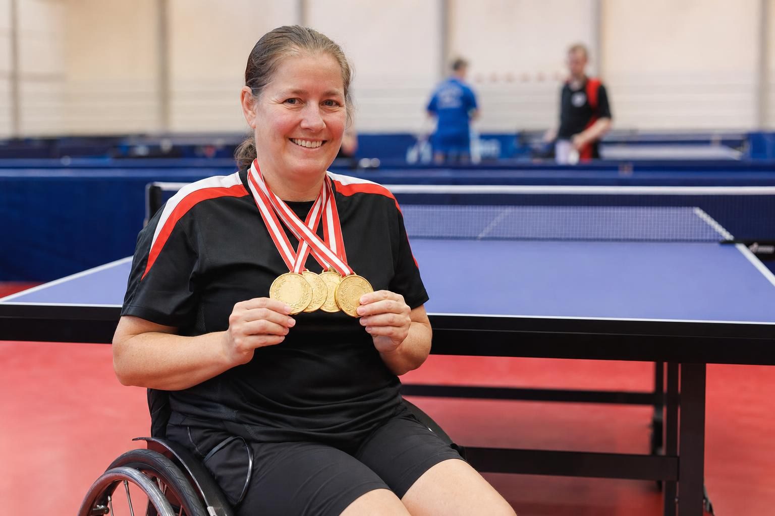 Eine lächelnde Frau im Rollstuhl hält vier Goldmedaillen vor Tischtennisplatten.