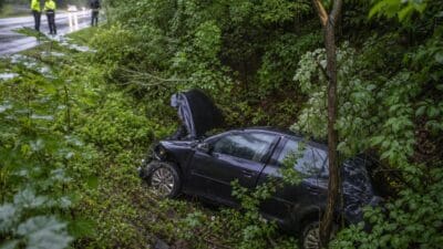 Beschädigter schwarzer Pkw mit offener Motorhaube in bewaldeter Böschung