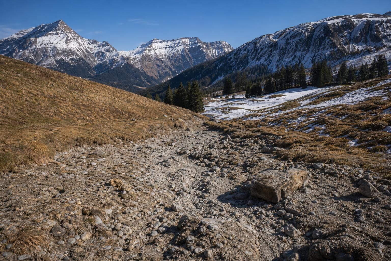 Trockener steiniger Pfad durch alpine Wiesen mit schneebedeckten Bergen im Hintergrund