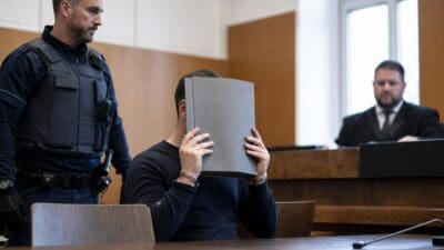 Ein Mann verdeckt sein Gesicht mit einem grauen Ordner in einem Gerichtssaal, neben ihm steht ein Polizist.