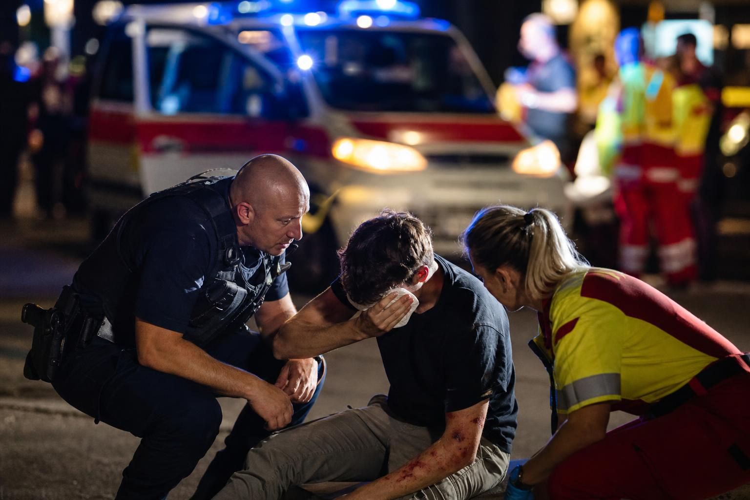 Polizist und Sanitäterin versorgen verletzten jungen Mann am Boden neben Rettungswagen bei Nacht