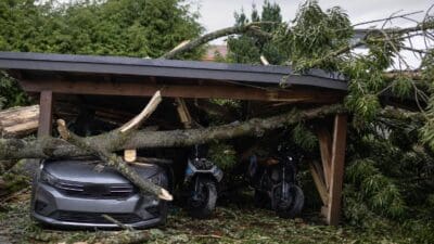 Graues Auto und zwei Motorräder unter einem von umgestürzten Bäumen zerstörten Carport mit Ästen und Trümmern auf dem Boden.