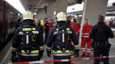 Zwei Feuerwehrleute mit Atemschutzgeräten stehen auf einem Bahnsteig hinter Absperrband