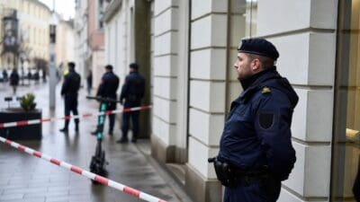 Polizist in Uniform steht vor Gebäude hinter rot-weißem Absperrband mit E-Scooter im Hintergrund