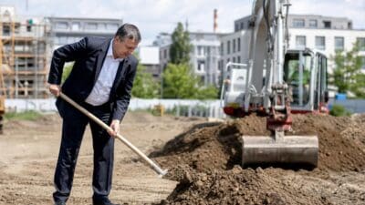Mann im dunklen Anzug hält Schaufel mit Erde auf Baustelle mit Bagger im Hintergrund