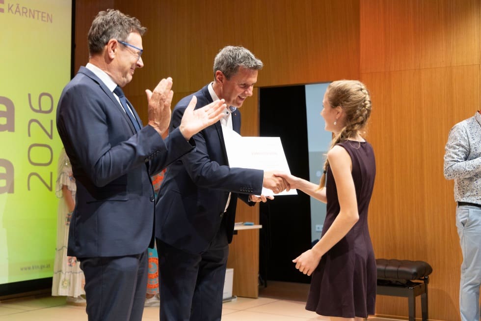 Eine junge Frau erhält eine Urkunde von einem Mann im Anzug während ein weiterer applaudiert