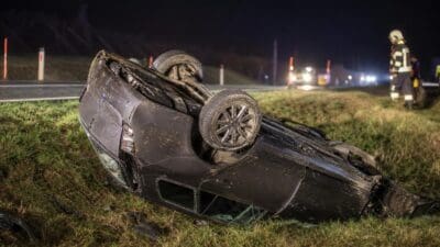 Überrolltes schwarzes Auto liegt nachts auf einer Wiese neben der Straße