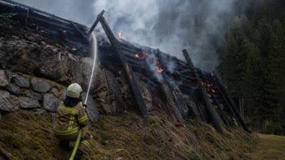 Feuerwehrmann in Schutzkleidung löscht brennende Holzverbauung auf steinigem Berghang