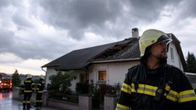 Feuerwehrmann in voller Schutzkleidung vor einem Haus mit zerstörtem Dach bei einem Einsatz