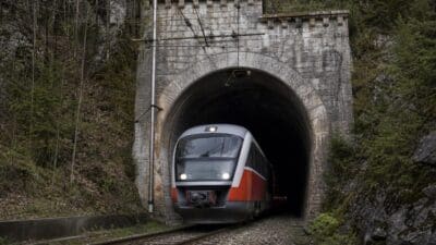 Roter und silberner Zug verlässt steinernes Tunnelportal in bewaldeter Berglandschaft