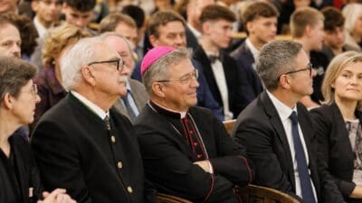 Bischof mit pinkem Birett sitzt neben einem Mann in Trachtenjacke bei einer Feier