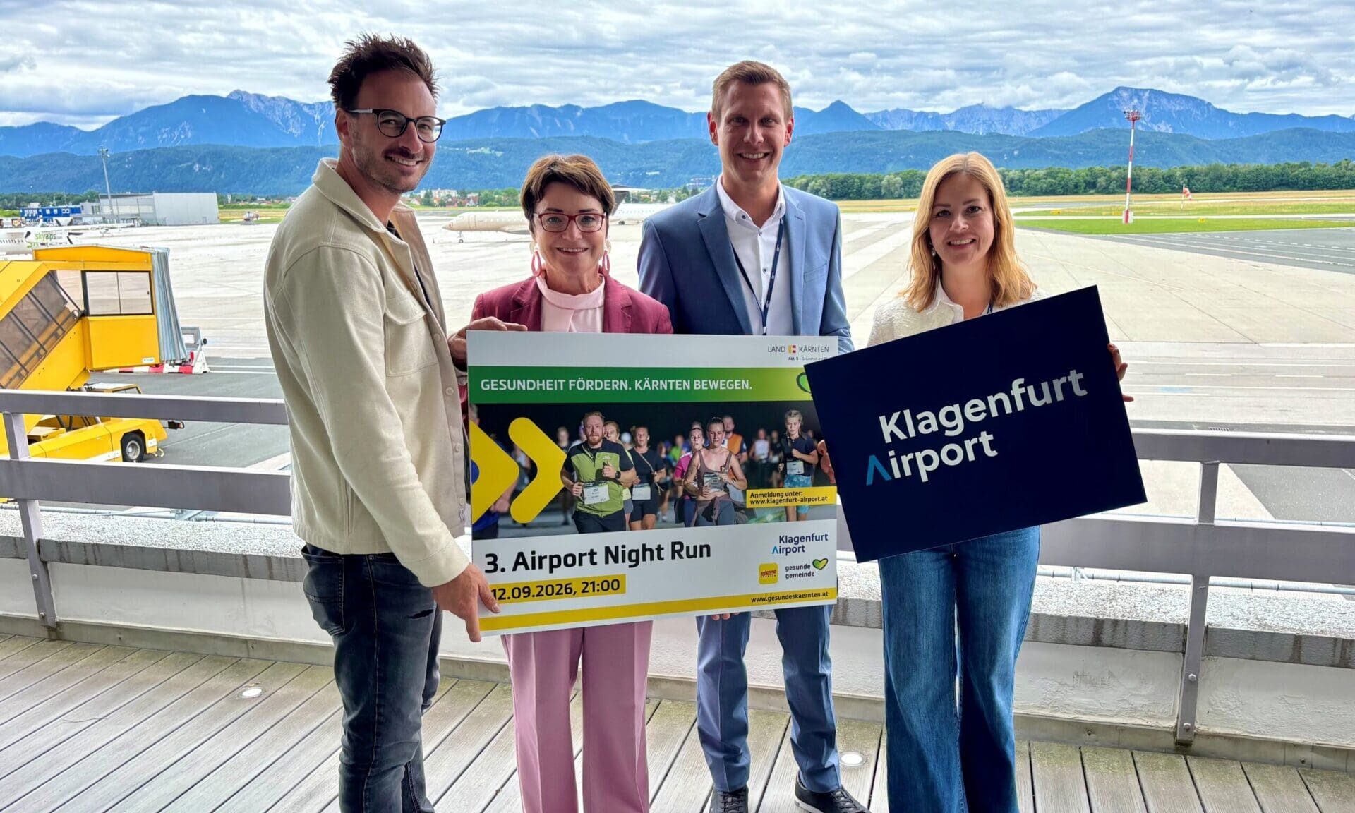 Gruppe von vier Personen mit Werbeschildern für den Airport Night Run vor der Kulisse des Flughafens Klagenfurt